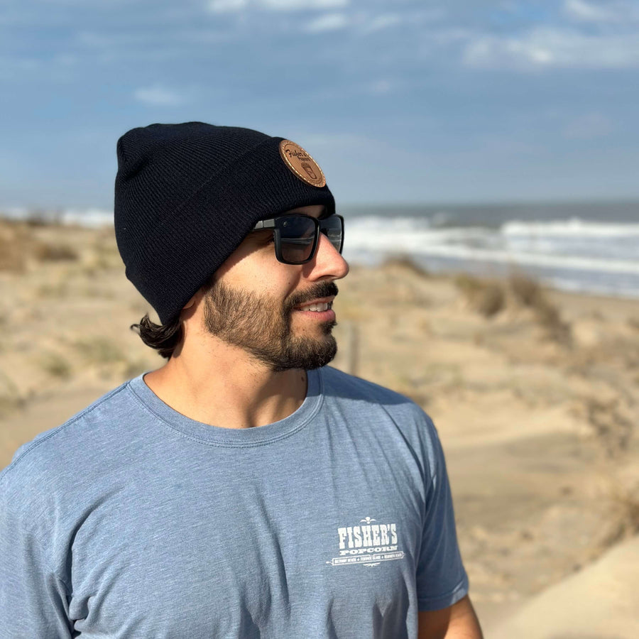 A man in a Fisher's Popcorn Fisher's Beanie and sunglasses, wearing a light blue t-shirt, stands on a sandy beach with the ocean and blue sky behind him.