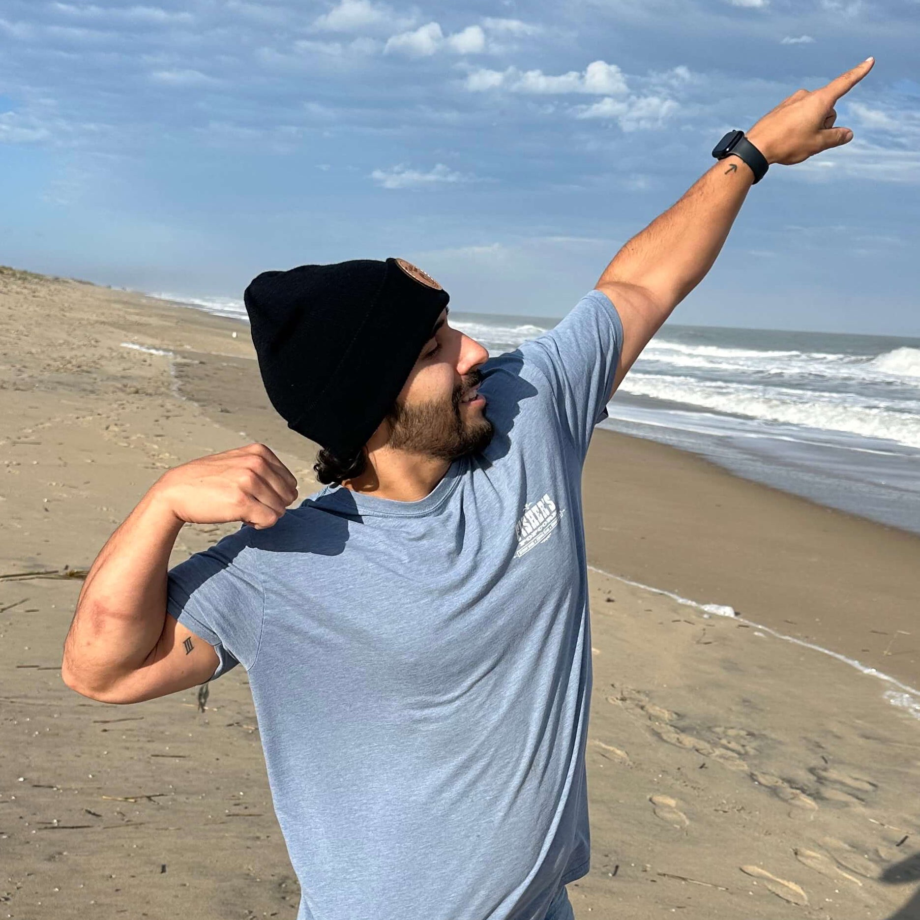 A man in a Fisher's Beanie by Fisher's Popcorn strikes a playful superhero pose on a sandy beach with waves and a blue sky—perfect for popcorn fans.