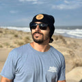 A bearded man in sunglasses, a Fisher's Popcorn Fisher's Beanie, and a light blue t-shirt stands on a sandy beach with grassy dunes and the ocean behind him under partly cloudy skies.