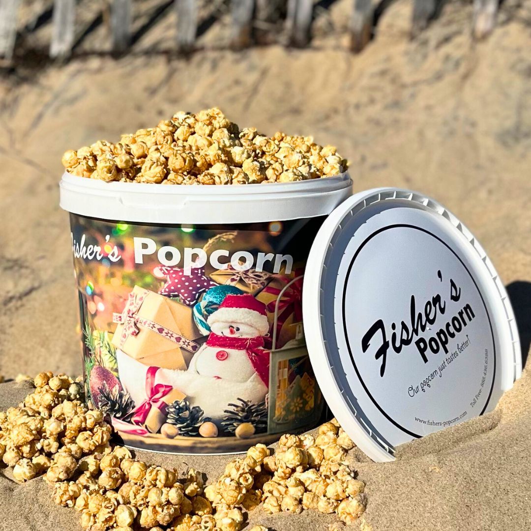 A Fisher's Popcorn Holiday Tub by Fisher's Popcorn, featuring a festive snowman and presents design, sits open on the sand with popcorn spilling out—perfect for sharing holiday popcorn gifts at the beach.