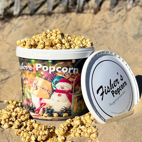 A Fisher's Popcorn Holiday Tub by Fisher's Popcorn, featuring a festive snowman and presents design, sits open on the sand with popcorn spilling out—perfect for sharing holiday popcorn gifts at the beach.