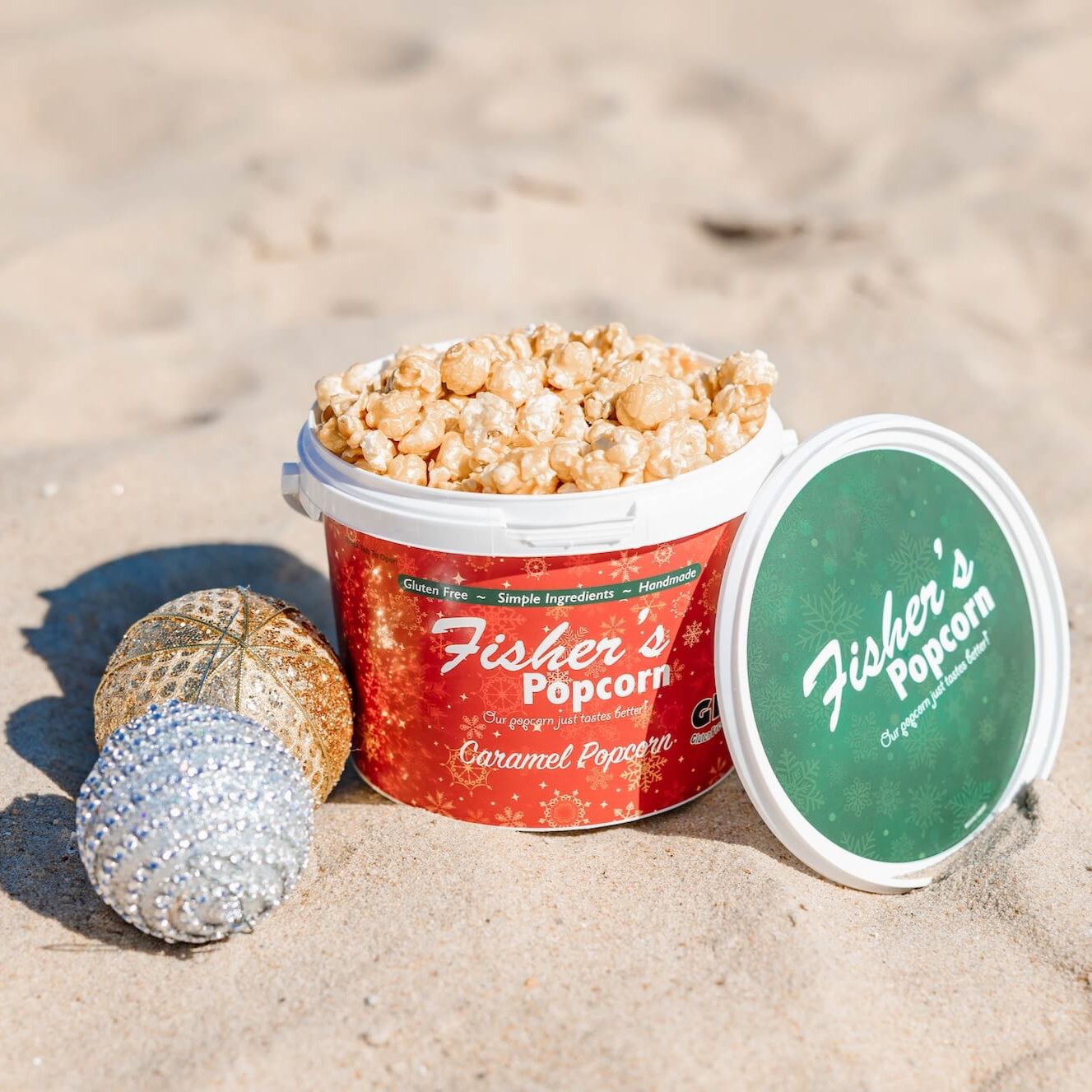 An open Fisher's Popcorn Holiday Tub rests on the sand, its caramel popcorn ready to enjoy. Gold and silver ornaments add festive flair—perfect inspiration for holiday gifts from Fisher's Popcorn.