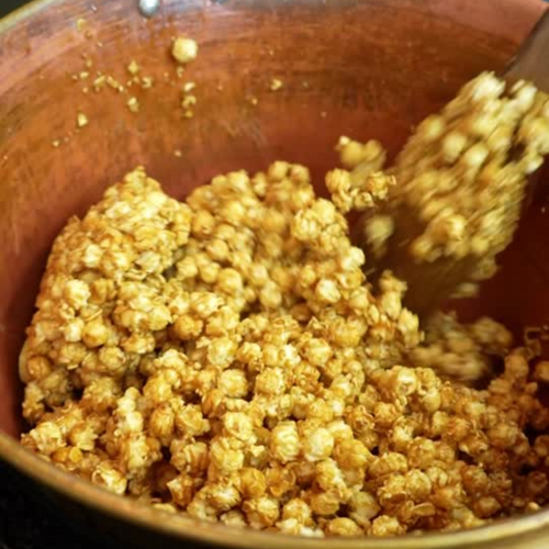 A close-up of Fisher’s Popcorn Tubs by Fisher's Popcorn shows golden brown caramel-coated clusters being stirred in a copper pot with a wooden spoon—just like in their classic caramel popcorn.