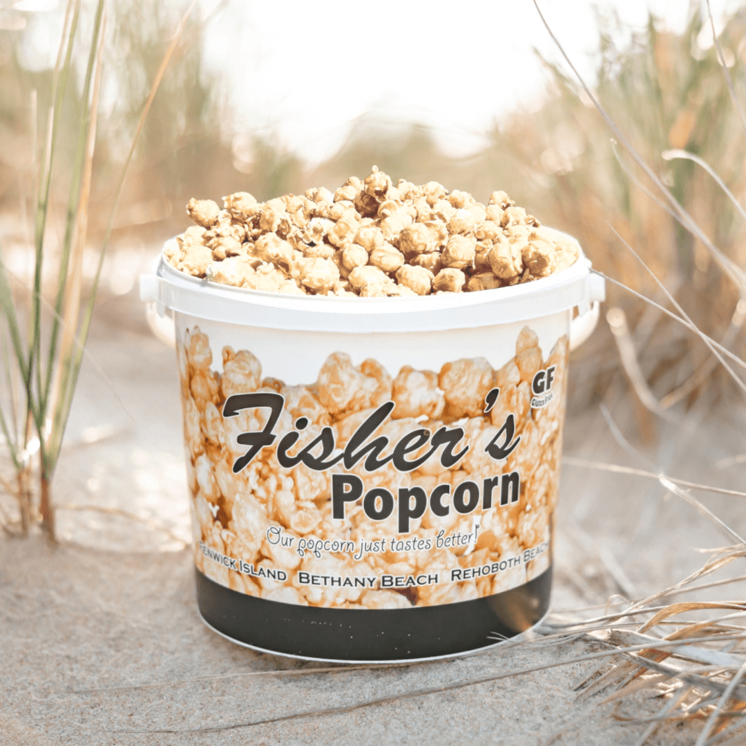 A Fisher's Popcorn Tub, filled with original caramel popcorn, sits on sandy ground amid beach grass. The branded bucket features images of popcorn and coastal scenes.