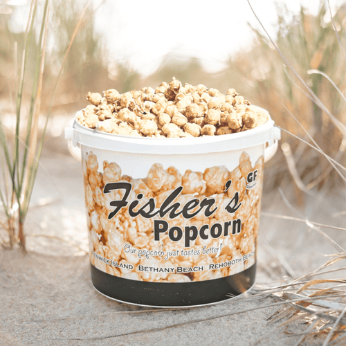 A Fisher's Popcorn Tub, filled with original caramel popcorn, sits on sandy ground amid beach grass. The branded bucket features images of popcorn and coastal scenes.