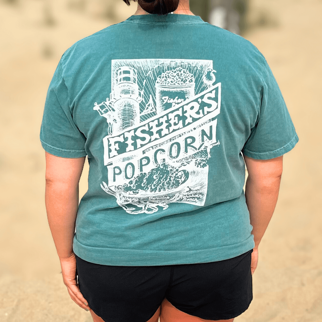 A person in a teal Fisher's Popcorn Fisher's T-Shirt featuring a white lighthouse and popcorn graphic, standing on sandy ground—the ideal beach shirt for popcorn fans.