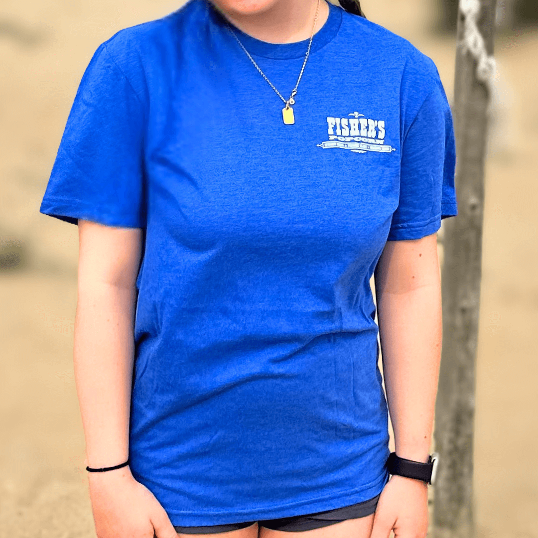 A person wearing a bright blue Fisher's T-Shirt from Fisher's Popcorn stands on sand outdoors. Ideal for popcorn lovers, their face is not visible.