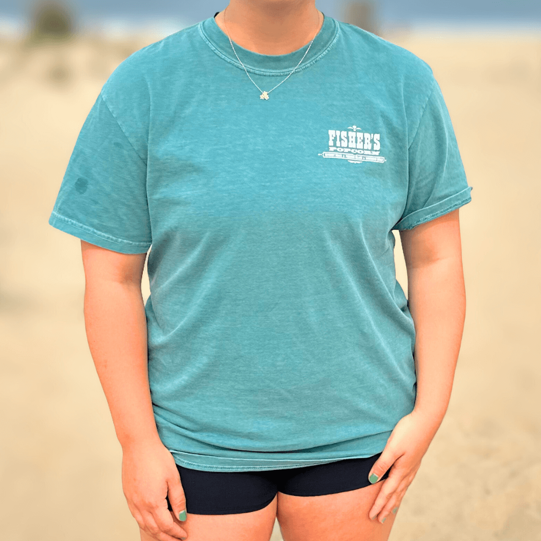 A person in a teal Fisher's Popcorn Fisher's T-Shirt and black shorts stands on a sandy beach, face not visible. Ideal for popcorn lovers who want the perfect beach shirt from Fisher's Popcorn.