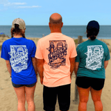 Three people stand on a sandy beach facing the ocean, each sporting a Fisher’s T-Shirt from Fisher’s Popcorn—a perfect day by the sea to show off your favorite beach shirt from this iconic popcorn brand.