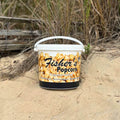 A large white Fisher's Popcorn Tubs container of caramel popcorn rests on sandy ground with beach grass and wooden planks, featuring "Bethany Beach, Rehoboth Beach, Ocean City" printed on the bucket. Brand: Fisher's Popcorn.
