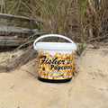 A large white Fisher’s Popcorn Tub sits on sandy ground with beach grass and a weathered wooden fence in the background, hinting at delicious caramel popcorn inside.