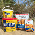 Old Bay Bags and Tubs from Fisher’s Popcorn are arranged on sand with grasses and dunes behind them, showcasing this sweet and savory treat in a tranquil beach setting.