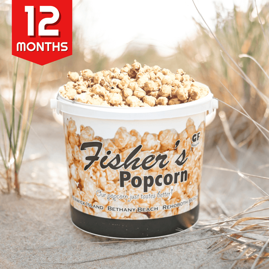 A large white tub of Fisher’s Popcorn sits on sandy ground with beach grass. A red “12 MONTHS” label in the corner highlights the Popcorn of the Month Club—a year-long subscription from Fisher’s Popcorn.