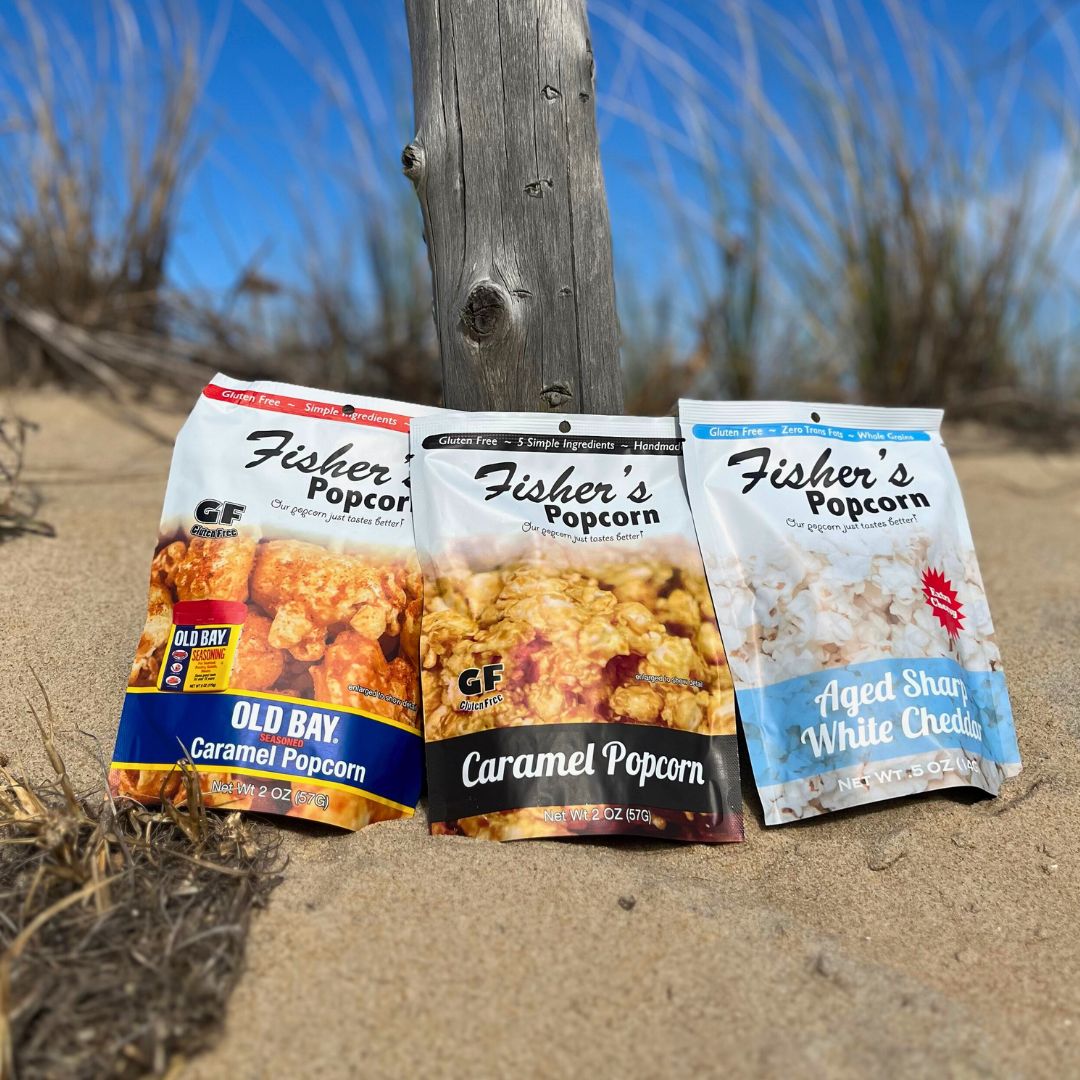 A Fisher's Popcorn Small Bag Sampler is propped in the sand by a wooden post, with dry grass and a bright blue sky in the background.