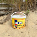 A Fisher's Popcorn Small Old Bay Tub rests on sandy ground outdoors, with a wooden fence and grass blurred in the background—a tasty addition to your beach day.