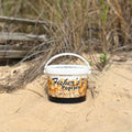 A Fisher’s Popcorn Tubs container from Fisher's Popcorn rests on sandy ground, with tall dry grass and weathered wooden planks in the background.