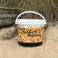 A Fisher's Popcorn Tubs container from Fisher's Popcorn rests on sandy ground near grass and wood, featuring original caramel popcorn on its label.