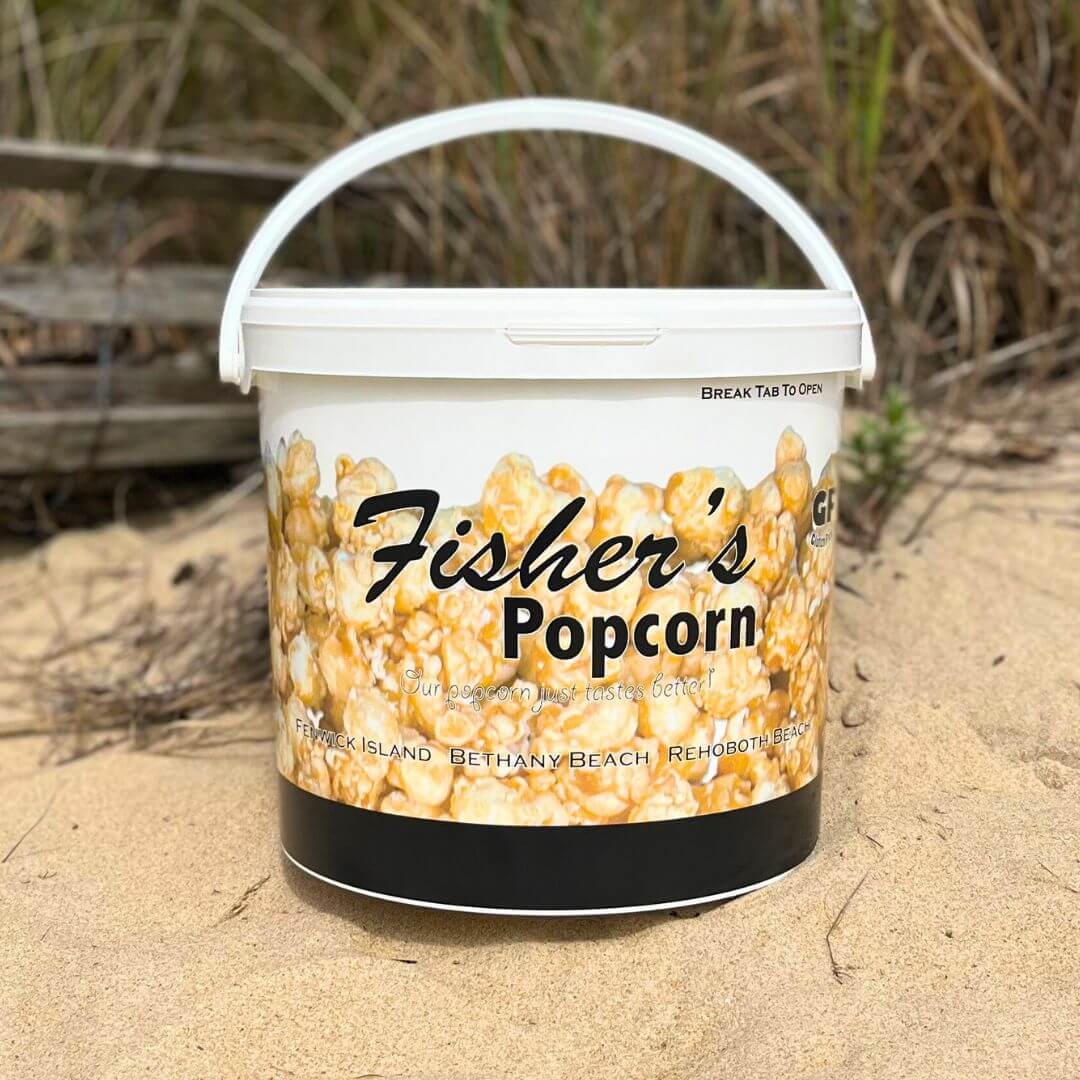 A large white Fisher’s Popcorn Tubs container rests on sand with dunes and wooden planks behind it. The tub showcases the brand's original caramel popcorn and lists beach town names along the bottom.