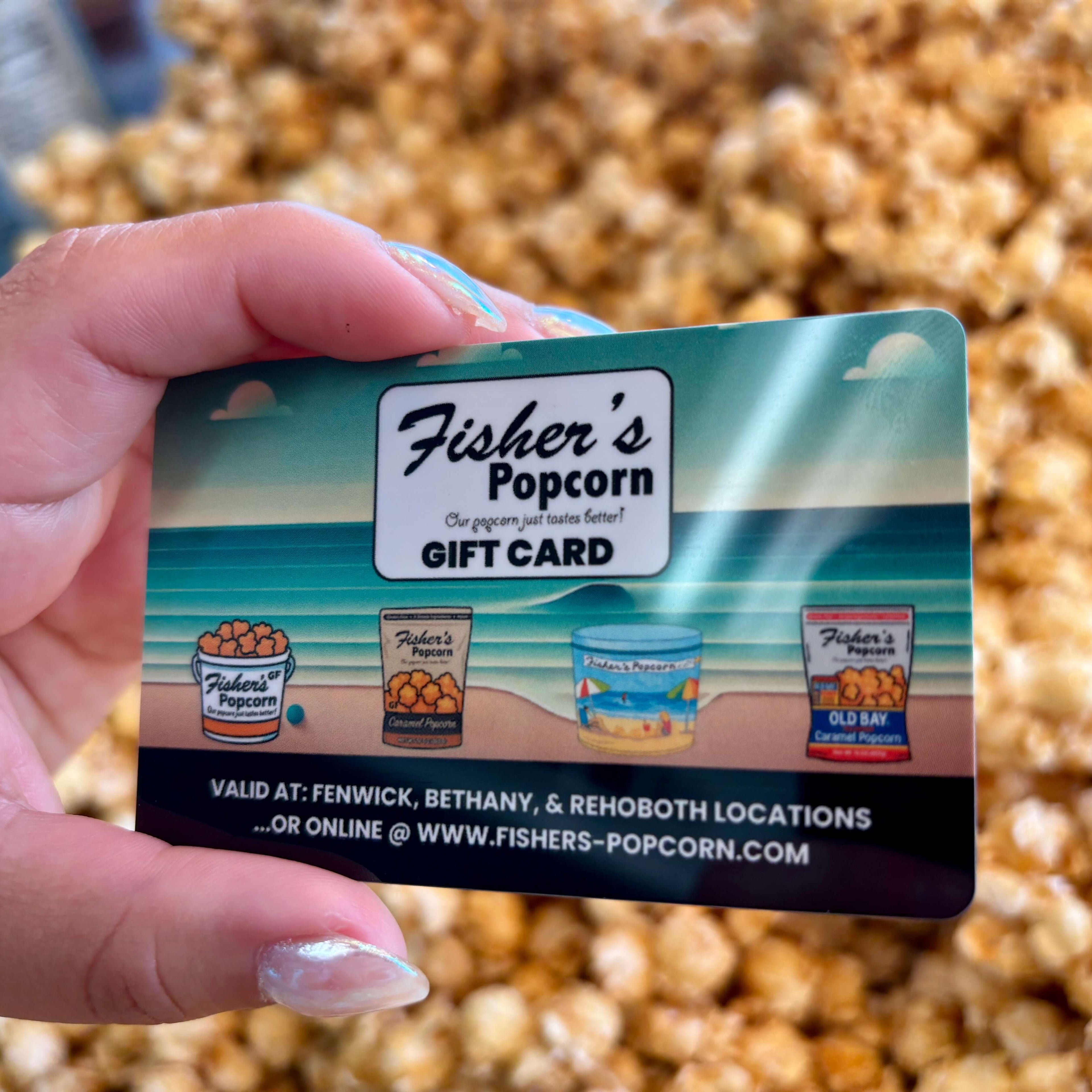 A hand holds a Fisher's Popcorn Gift Card (Physical) by Loopz Gift Cards, featuring an ocean scene, images of popcorn tubs, store locations, and the website in front of a pile of popcorn.