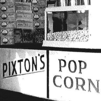 A black-and-white photo of a popcorn stand with signs reading PIXTONS and POP CORN. A popcorn machine filled with popcorn is visible on the counter.