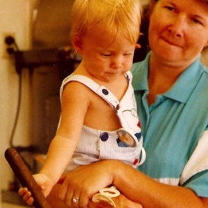 A woman in a teal and white shirt holds a small blond child wearing light-colored overalls with one strap unfastened. The child looks down, focusing on something out of view.
