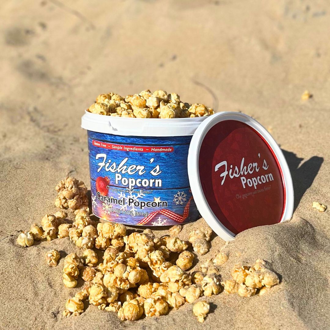 A tub of Fishers Caramel Popcorn sits open on sandy ground, with popcorn spilling out around it. The red lid lies to the right of the tub. The scene is brightly lit by sunlight.