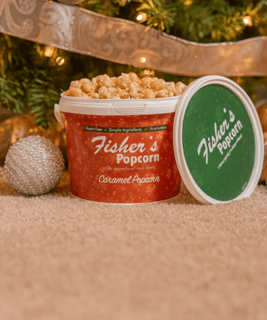 A tub of Fishers Caramel Popcorn sits on a carpet in front of a decorated Christmas tree, with its green lid resting nearby and a silver ornament beside it.