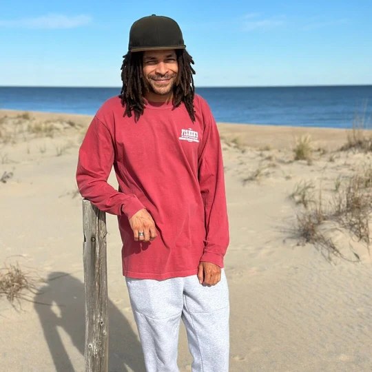 Man in a red Fisher's long sleeve tee leaning on a dune fence post at the beach